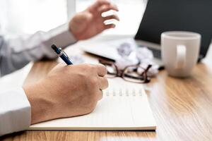 une homme utilisation crayon écrire sur carnet papier et froisser papier des lunettes café tasse avec ordinateur pour Bureau travail photo