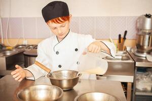 roux garçon dans du chef costume cuisiniers Crêpes dans cuisine, pétrit pâte, assistant fait du petit déjeuner. verser Lait dans bol photo