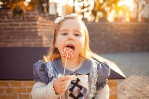 magnifique peu fille mange, lèche avec langue délicieux bonbons, habillé comme sorcière, en plein air, automne, octobre 31, content Halloween. portrait drôle. photo