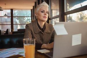 Sénior femme de 50 ans est magnifique et élégant, est assis dans café, travaux avec ordinateur portable, free-lance designer. regards super, content vie, actif mode de vie photo