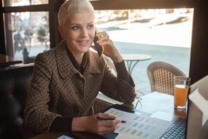 Sénior femme de 50 ans est magnifique et élégant, est assis dans café, travaux avec ordinateur portable, free-lance designer. regards super, content vie, actif mode de vie. téléphone intelligent. photo