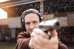 une homme pousse une pistolet, visée à le cible. une homme portant protecteur écouteurs. une mur et une toit avec balle des trous. Extérieur tournage intervalle proche en haut photo