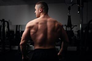 portrait de une magnifique Masculin athlète bodybuilder dans salle de sport, faible clé, obscurité. poser, entraîneur. retour photo