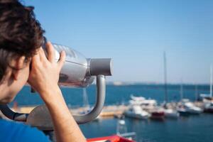 homme regards par Stationnaire jumelles. binoscope sur observation plate-forme près mer. port de mer dans Odessa. coûteux yachts sur le jetée. commercial machine, vente entreprise. proche en haut photo