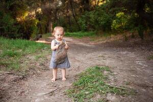 une enfant recueille des ordures dans le forêt, une peu fille porte une Plastique bouteille contre une Contexte de magnifique des arbres. le enfant a trouvé une des ordures pouvez dans le les bois. protéger le environnement photo