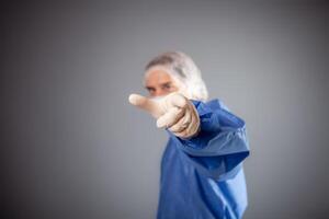 une Beau Masculin chirurgien est une médecin sur une gris Contexte dans le studio. une homme dans une peignoir, une médical masque, une chapeau. le la personne points le sien doigt à le caméra, appels à joindre quelque chose ou refuser. photo