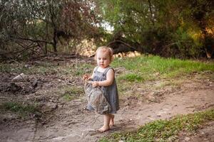 une enfant recueille des ordures dans le forêt, une peu fille porte une Plastique bouteille contre une Contexte de magnifique des arbres. le enfant a trouvé une des ordures pouvez dans le les bois. protéger le environnement photo