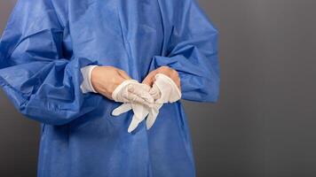 une femme médecin prend de sa blanc gants. une femme dans une bleu chirurgical peignoir, une gris arrière-plan, un vide endroit photo