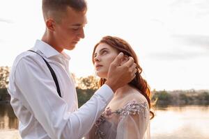 une magnifique mariage couple sur le berge de rivière à le coucher du soleil. une femme dans une gris robe avec fleurs, une homme dans une costume et bretelles étreinte chaque autre, l'amour. les amoureux sur une marcher photo