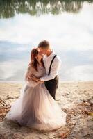 une magnifique mariage couple sur le berge de rivière à le coucher du soleil. une femme dans une gris robe avec fleurs, une homme dans une costume et bretelles étreinte chaque autre, l'amour. les amoureux sur une marcher photo