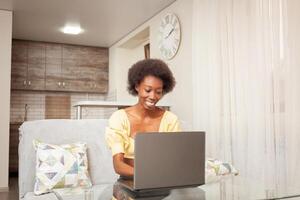 une africain américain femme pigiste, travail de maison. femme dans de face de portable est content, souriant. gagnant loterie, embauché, les stocks avoir ressuscité, crypto-monnaie a grandi, bien nouvelles. photo