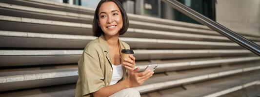 content Urbain fille les boissons sa à emporter café et parchemins alimentation sur téléphone intelligent. asiatique femme est assis sur escaliers avec thé et en portant mobile téléphone photo