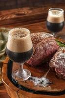 deux verres de tulipe en sueur de bière pression noire avec contre-filet grillé sur une planche à découper en bois. table en bois. photo