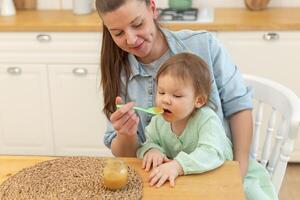 content famille à maison. mère alimentation sa bébé fille de cuillère dans cuisine. peu bambin enfant avec désordonné marrant visage mange en bonne santé nourriture à maison. Jeune femme maman donnant nourriture à enfant fille photo