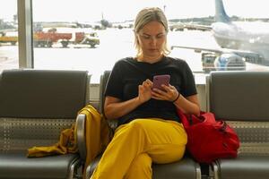 aéroport Terminal. femme attendre pour vol en utilisant téléphone intelligent. fille avec cellule téléphone dans aéroport surfant l'Internet social médias applications. en voyageant femelle dans embarquement salon de Compagnie aérienne moyeu. en voyageant fille photo