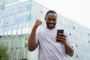 content africain américain homme euphorique gagnant avec téléphone intelligent sur rue dans ville. la personne gars à la recherche à cellule téléphone en train de lire génial nouvelles avoir bien résultat gagnant en ligne offre sentiment étonné. gagnant geste photo