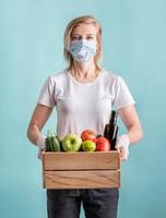 femme blonde dans un masque et des gants tenant une boîte en bois pleine de légumes photo