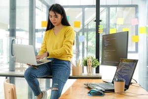 une programmeuse asiatique porte une chemise jaune, s'assoit sur des étagères et regarde l'écran d'ordinateur portable sur sa jambe. photo