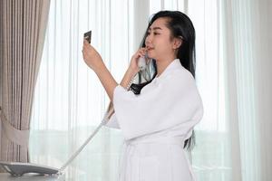 une belle jeune fille en peignoir dans un peignoir tenant une carte de crédit tout en parlant au téléphone dans la chambre d'hôtel photo