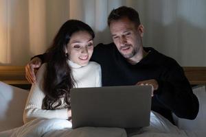 couple cuacasian regardant des films du programme streming dans l'ordinateur ensemble sur le lit à la maison dans la nuit photo