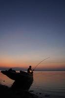 au bord du lac, un pêcheur asiatique assis sur un bateau et utilisant une canne à pêche pour attraper du poisson au lever du soleil photo