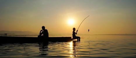au bord du lac, un pêcheur asiatique assis sur un bateau tandis que son fils se tient debout et utilise une canne à pêche pour attraper du poisson au lever du soleil photo
