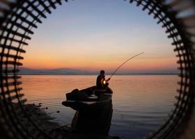 au bord du lac, un pêcheur asiatique assis sur un bateau et utilisant une canne à pêche pour attraper du poisson au lever du soleil photo