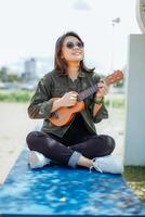 en jouant ukulélé de Jeune magnifique asiatique femme portant veste et noir jeans posant en plein air photo