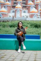 en jouant ukulélé de Jeune magnifique asiatique femme portant veste et noir jeans posant en plein air photo