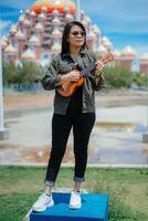 en jouant ukulélé de Jeune magnifique asiatique femme portant veste et noir jeans posant en plein air photo