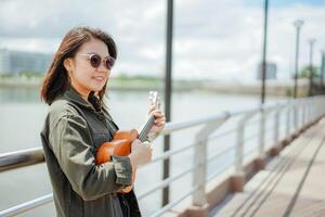 en jouant ukulélé de Jeune magnifique asiatique femme portant veste et noir jeans posant en plein air photo