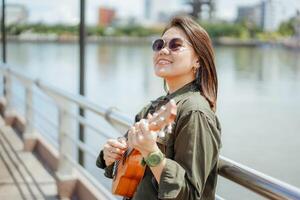 en jouant ukulélé de Jeune magnifique asiatique femme portant veste et noir jeans posant en plein air photo