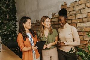 Trois Jeune affaires femmes avec numérique tablette permanent par le brique mur dans le industriel style Bureau photo