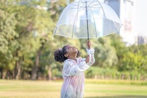 de bonne humeur enfant fille avec un parapluie en jouant avec pluie dans le parc, mignonne peu enfant fille en jouant en plein air dans le jardin photo