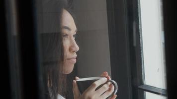 Jeune métisse femme marche tenant tasse à deux mains, se dirige vers la fenêtre, en prenant une gorgée, regardant à travers la fenêtre de manière contemplative photo