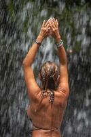 magnifique Jeune femme se détend en dessous de une cascade. sexy fille dans une bikini posant près une cascade dans le tropiques. une voyageur baignades en dessous de ruisseaux de l'eau écoulement de le montagnes. photo