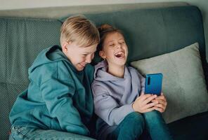deux les enfants séance sur une canapé en riant tandis que en utilisant une cellule téléphone photo