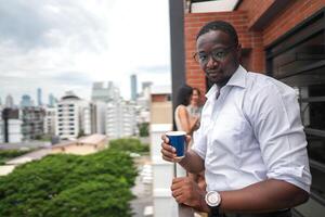 africain affaires intelligent homme des stands à Extérieur terrasse bâtiment. gens avec sur main dans bien sentiment avec ville espace bâtiment. relaxant de gens affaires Matin. photo