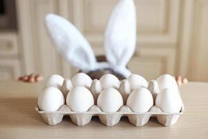 non visage mignonne enfant avec lapin oreilles est cache en dessous de le tableau. blanc biologique poulet des œufs dans une artisanat boîte supporter sur le table dans le cuisine. Pâques concept. photo