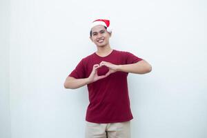 Jeune asiatique homme portant une Père Noël claus chapeau exprimer l'amour par formant une cœur symbole avec le sien mains isolé par une blanc Contexte pour visuel la communication photo