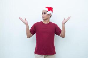 Jeune asiatique homme portant une Père Noël claus chapeau souriant, choc et montrer du doigt à sa côté isolé par blanc Contexte pour visuel la communication photo