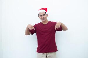 Jeune asiatique homme portant une Père Noël claus chapeau souriant, choc et montrer du doigt à sa côté isolé par blanc Contexte pour visuel la communication photo