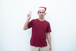 Jeune asiatique homme portant une Père Noël claus chapeau souriant, choc et montrer du doigt à sa côté isolé par blanc Contexte pour visuel la communication photo