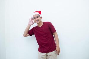 Jeune asiatique homme portant une Père Noël claus chapeau exprimer écoute avec main à oreille isolé par une blanc Contexte pour visuel la communication photo