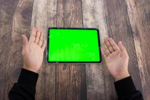 moquer en haut de une main en portant un iPad tablette avec une écran vert contre une en bois texture Contexte photo