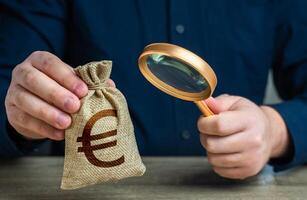 une homme examine une euro argent sac avec une grossissant verre. vérification le origine de capital. anti argent blanchissage, impôt évasion. étude termes et conditions sur dépôt ou prêt. trouver investissements photo