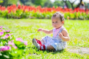 bébé garçon en jouant dans ensoleillé fleur jardin. adorable fils bébé avec une pensif expression est assis dans une ensoleillé jardin, entouré par verdure et vibrant rose et rouge fleurit. enfant vieilli 1 année vieux. photo