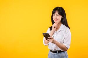 portrait magnifique asiatique Jeune femme portant blanc chemise et denim les plantes réfléchi corps geste avec téléphone intelligent isoler sur Jaune studio Contexte. en pensant positif dans en ligne nouvelles concept. photo