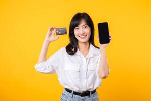 portrait asiatique Jeune femme portant blanc chemise et denim jean avec content sourire en portant crédit carte à payant achats en ligne sur téléphone intelligent isolé sur Jaune Contexte. achats en ligne concept. photo