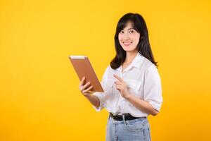 Jeune asiatique femme portant blanc chemise sourit vivement tandis que en portant numérique tablette contre Jaune arrière-plan, représente une sur de soi entreprise ouvrier embrassement moderne La technologie concepts. photo